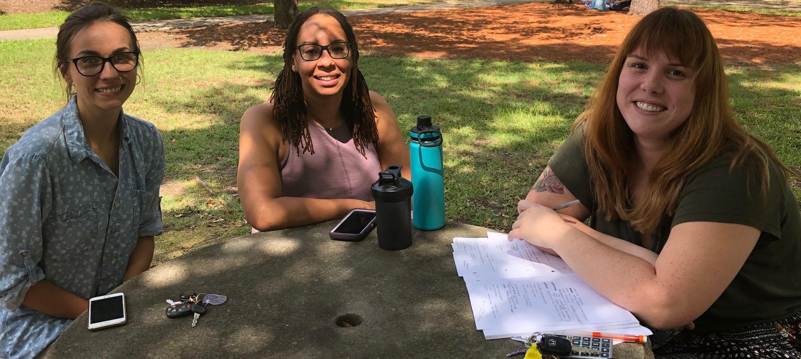 Three smiling, female, 白人学生围坐在一起, grey stone table: the first has brown hair with blond highlights worn down and is wearing a black shirt and a black skirt with multi-colored polka dots and is pictured with a stack of paper under her crossed arms on the table with a ring of keys including yellow and white cards and a black cell phone; the second has brown hair worn down and black rimmed glasses with a lavendar pink shirt, she is pictured with a tall, teal and black water bottle, 一个较短的黑色固体水瓶, and her cell phone with a lavendar back and white edges; and the third has on a blue, 白色圆点领衬衫和棕色裤子, 照片中她拿着车钥匙和白色的手机, 面部上下部分的横杠.